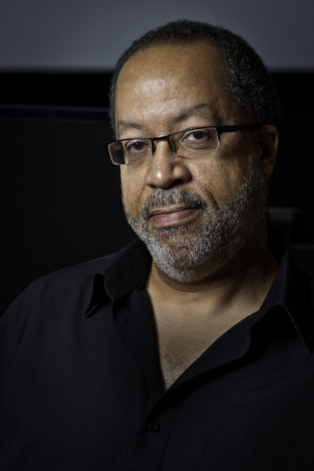 A black man with gray beard and short black hair wearing rectangular framed glasses and an open necked button down black shirt